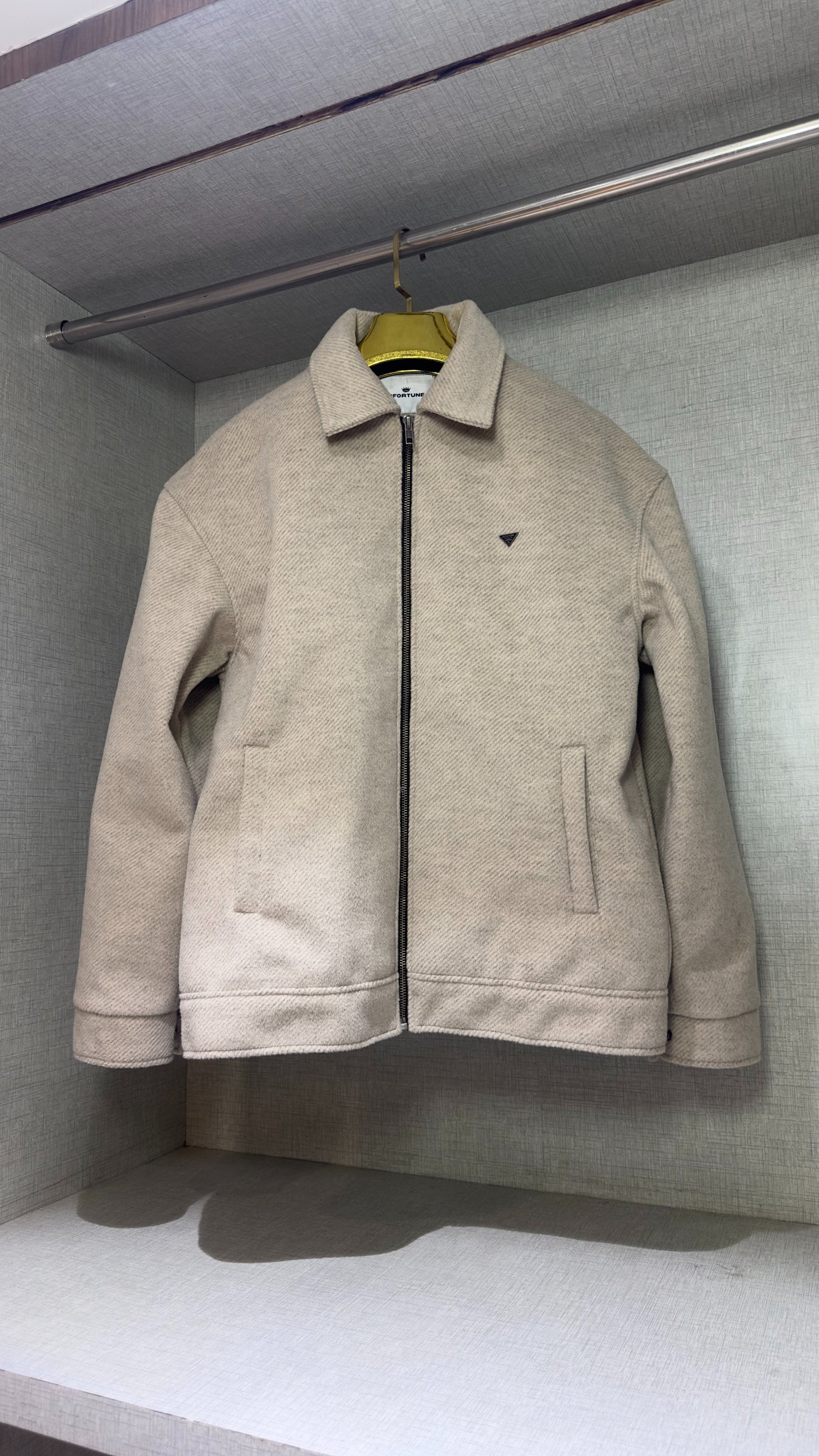 The full-front view of a cream-colored tweed jacket displayed in a wardrobe niche, its boxy shoulders and straight-cut sleeves fully visible. The jacket’s structured silhouette casts a soft shadow across the bottom shelf, highlighting the contrast between its textured surface and the smooth fabric lining of the closet.