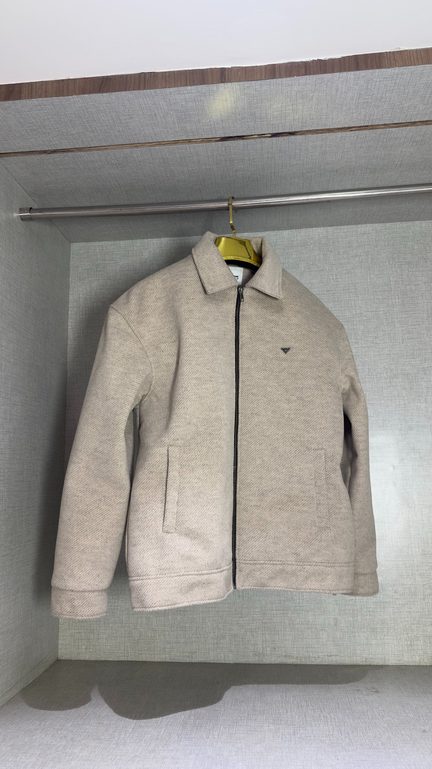 A cream tweed jacket with a boxy, oversized cut hangs alone on a sleek gold hanger inside a softly lit wardrobe alcove. Its straight hem and welt pockets sit symmetrically on either side of the central black zipper, while the garment’s nubby tweed fabric catches the light to reveal a subtle herringbone pattern.