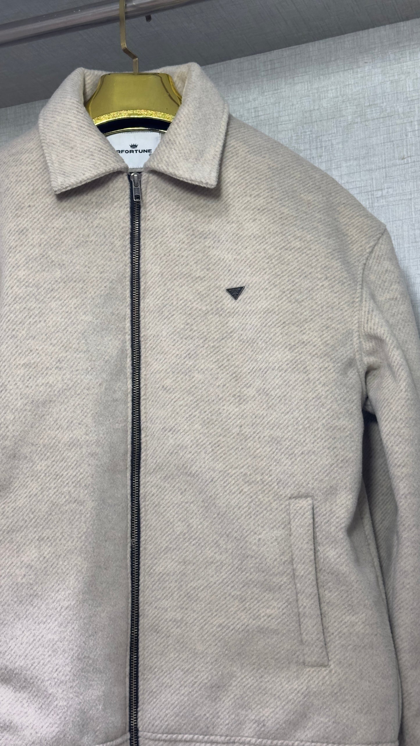 A close-up of the upper torso of a cream tweed jacket, emphasizing its pointed collar and exposed black-metal zipper. A small triangular logo patch adorns the left chest, and the dense, textured weave of the fabric is rendered in fine detail against the pale gray backdrop of the wardrobe lining.