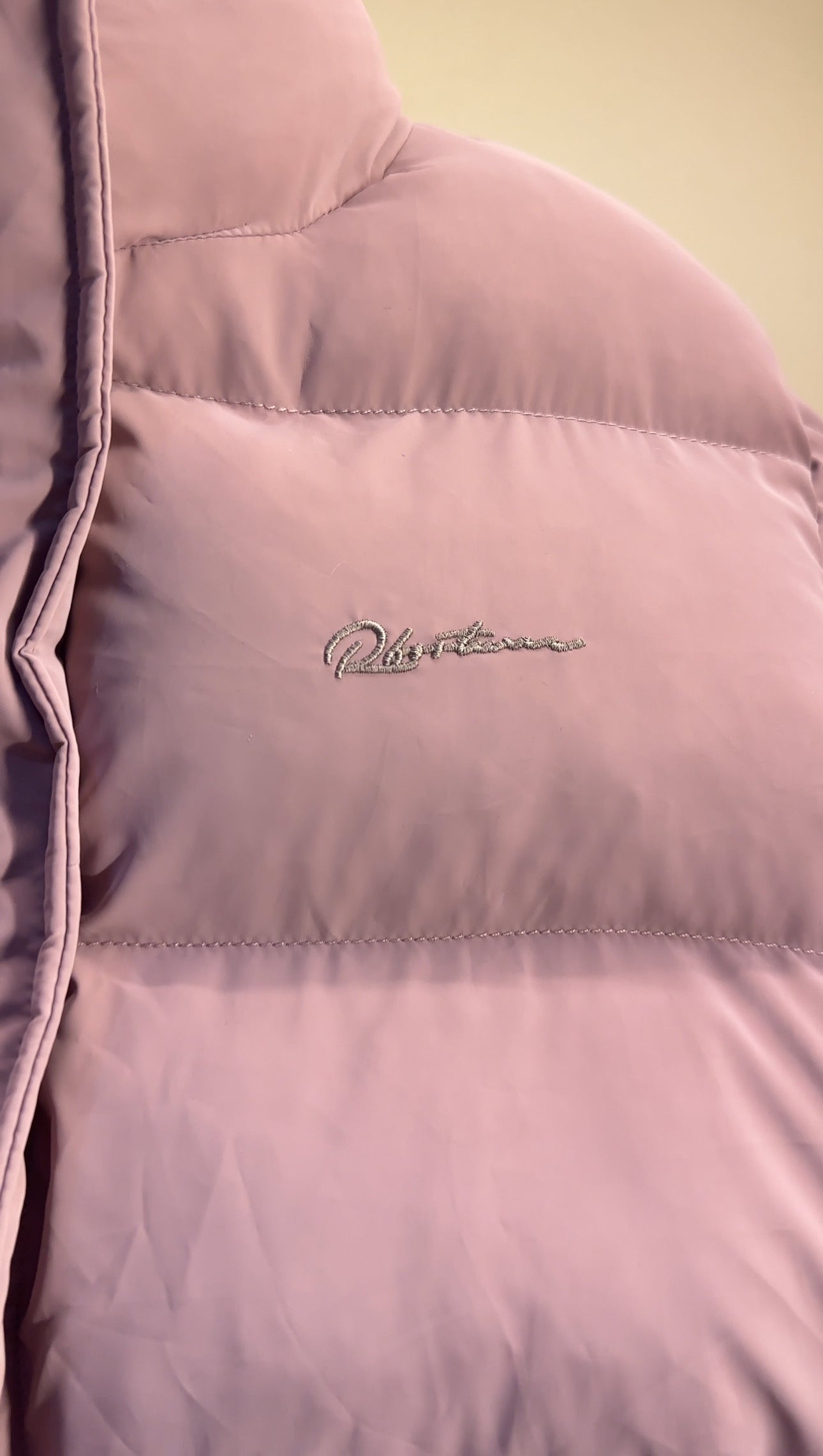 Close-up of the chest embroidery on a light pink Puffer jacket women wear, showing delicate stitched branding on the soft quilted fabric.
