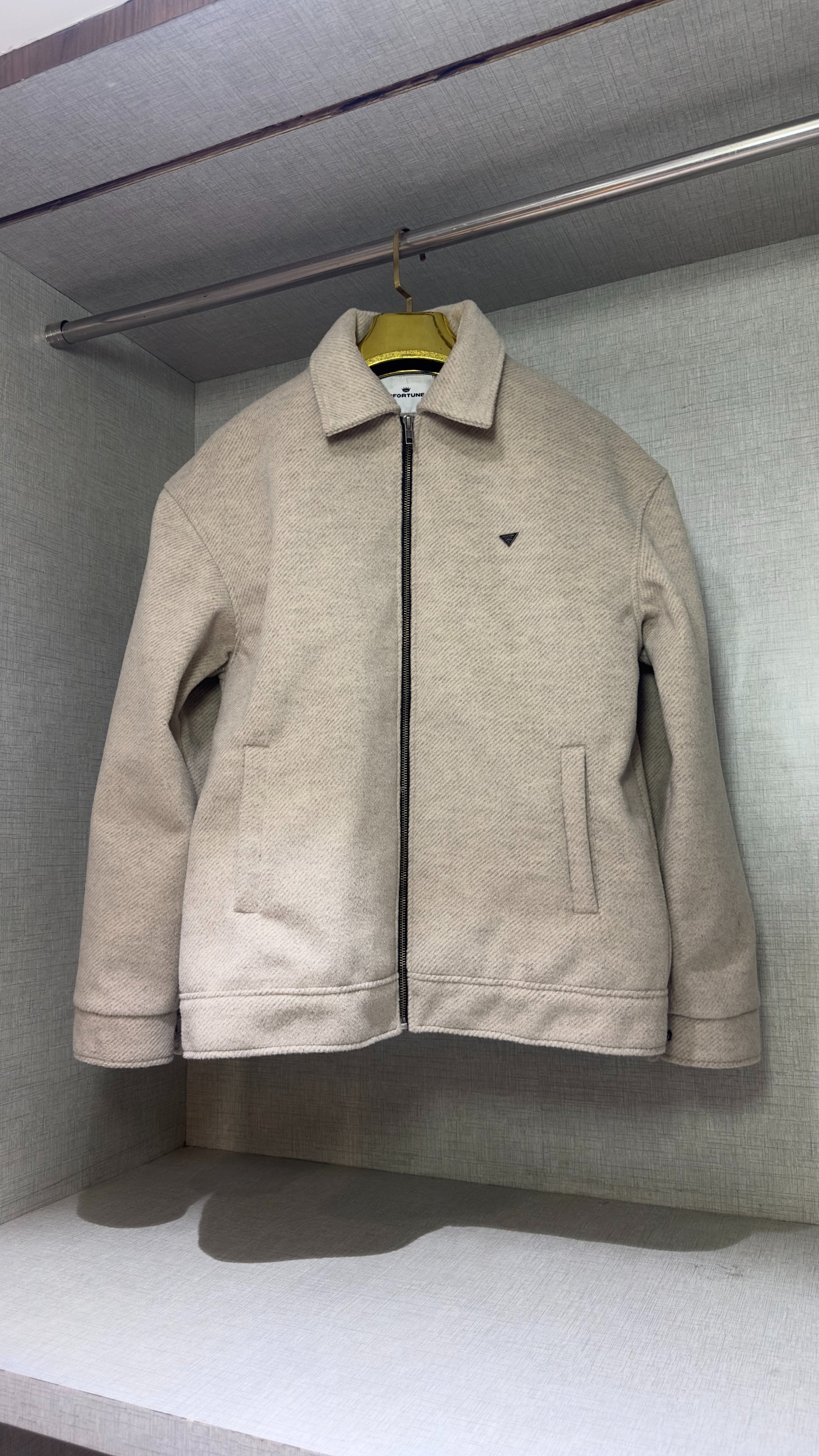 The full-front view of a cream-colored tweed jacket displayed in a wardrobe niche, its boxy shoulders and straight-cut sleeves fully visible. The jacket’s structured silhouette casts a soft shadow across the bottom shelf, highlighting the contrast between its textured surface and the smooth fabric lining of the closet.