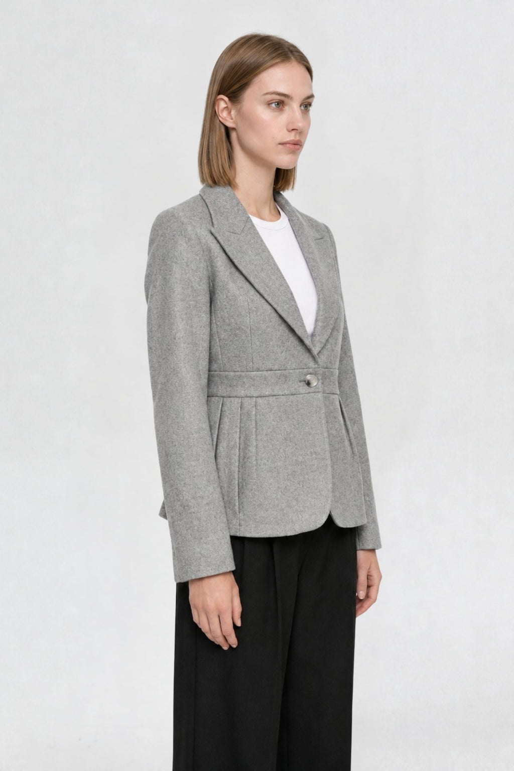 Side view of grey short blazer coat showing peaked lapel, single gold button, and pleated waist seam