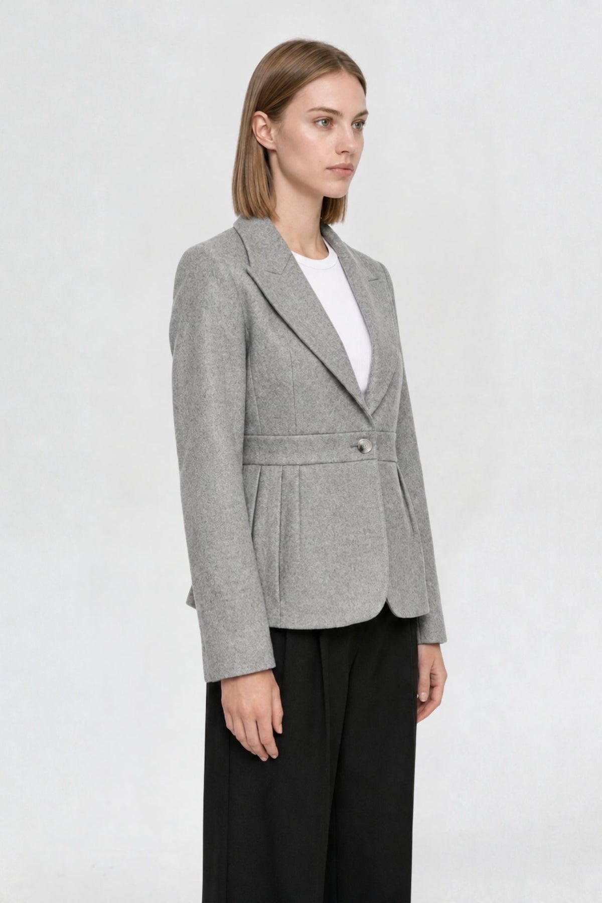 Side view of grey short blazer coat showing peaked lapel, single gold button, and pleated waist seam